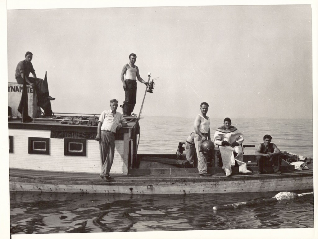 The Boston salvage crew that blew up the hull of the SS City of Salisbury after it sank in the fall of 1938. The hull was a navigation hazard. See the "Dynamite" and "Explosives" signs on the cabin at left. At right, the diver is seated in his white dive suit, with his companion, standing, holding the bronze diving helmet.